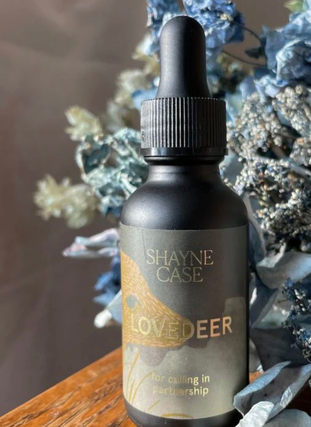 Bottle of Shaye Case Lovedeer on a wooden surface with decorative plants in the background
