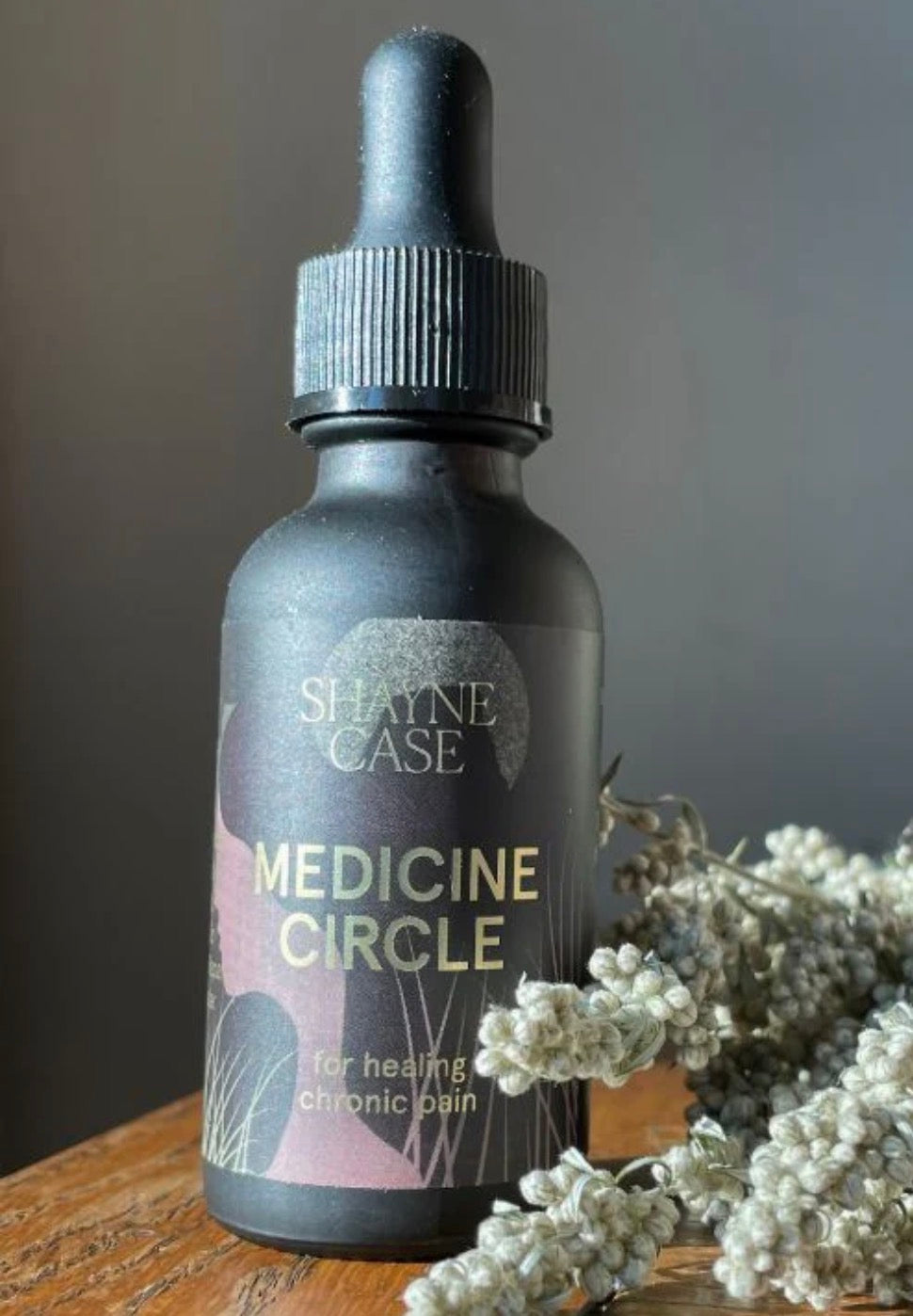 Bottle labeled 'Shayne Case Medicine Circle' with dried flowers on a wooden surface.